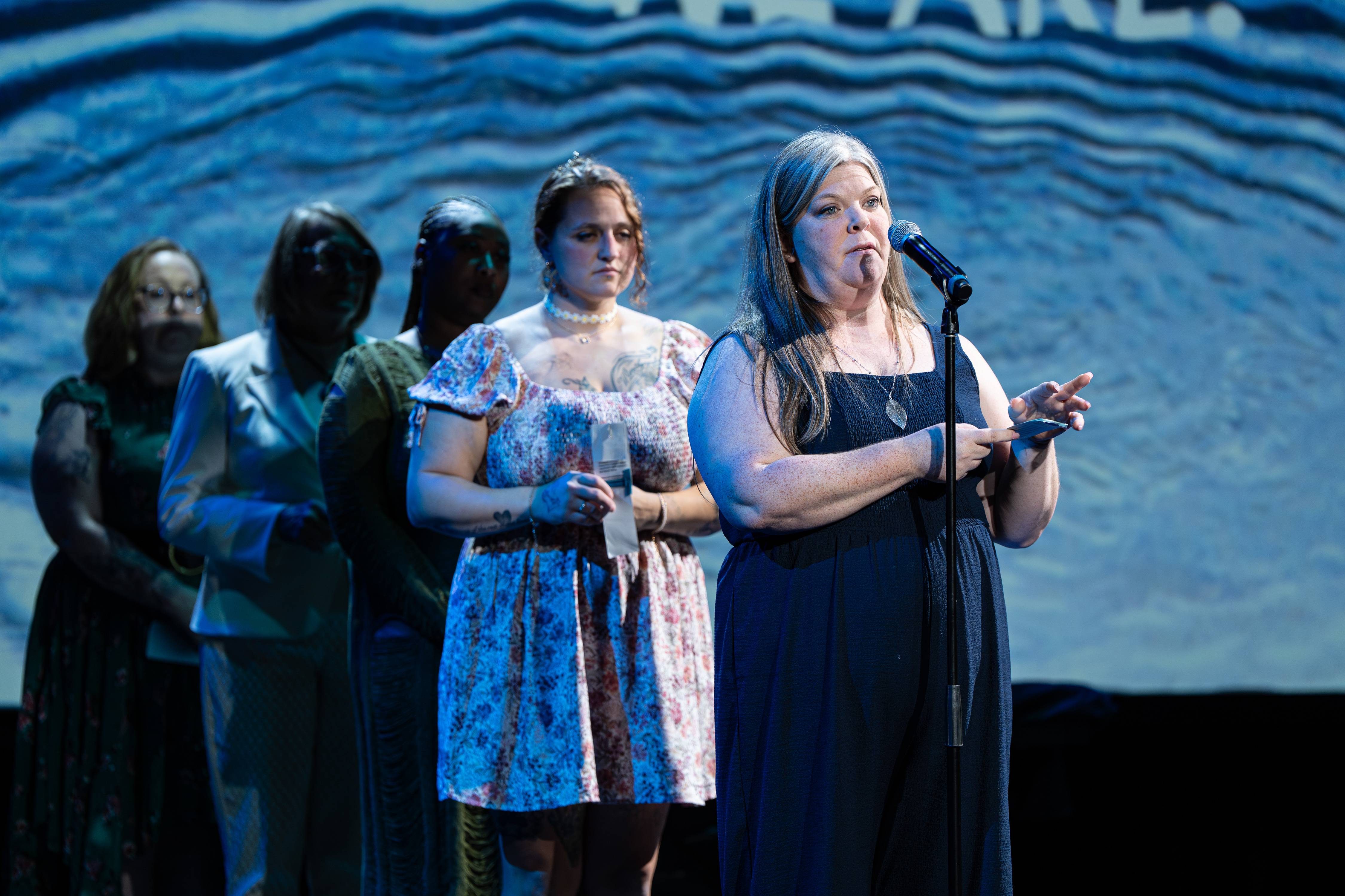 Five women stand in a row on stage in front of a blue wave-patterned backdrop reading 'WE ARE'; a woman in a navy dress speaks into a microphone in the foreground.