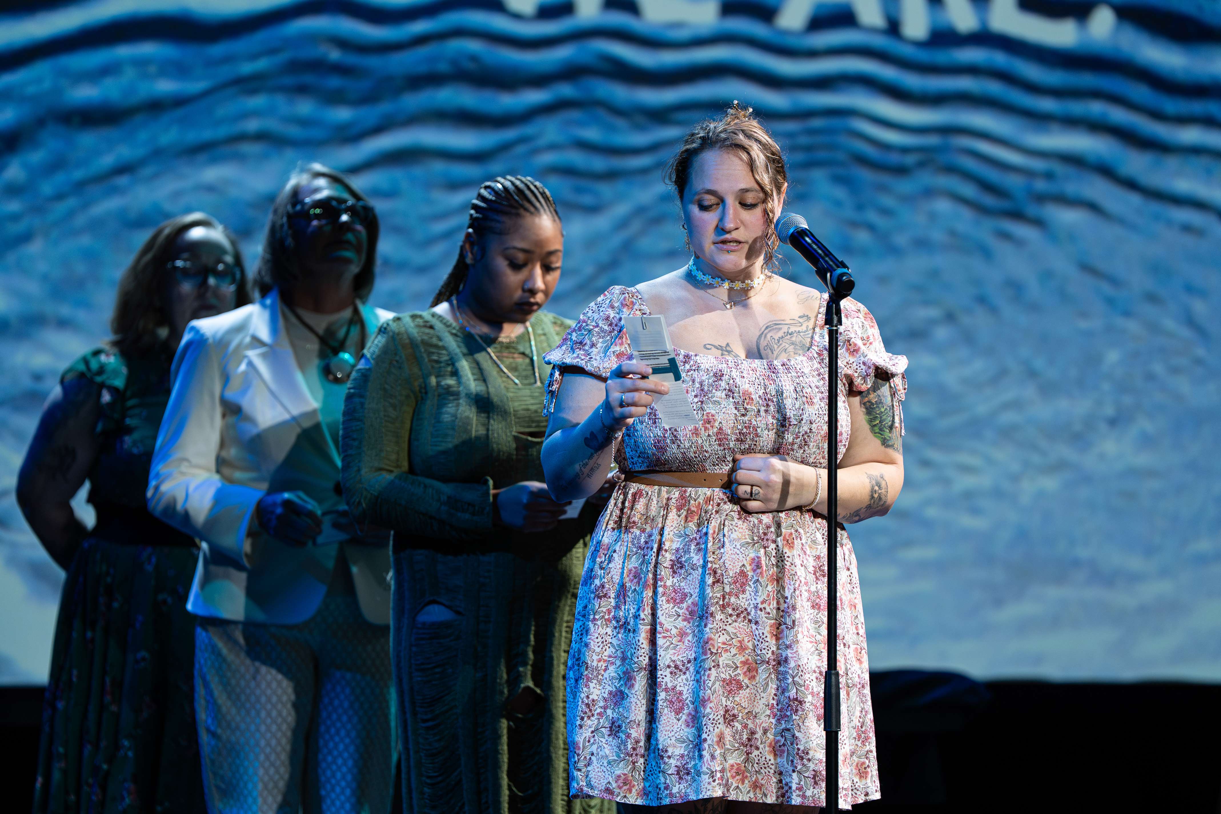 A young woman in a floral dress and daisy choker reads from a card into a microphone, with three other women standing behind her on stage.