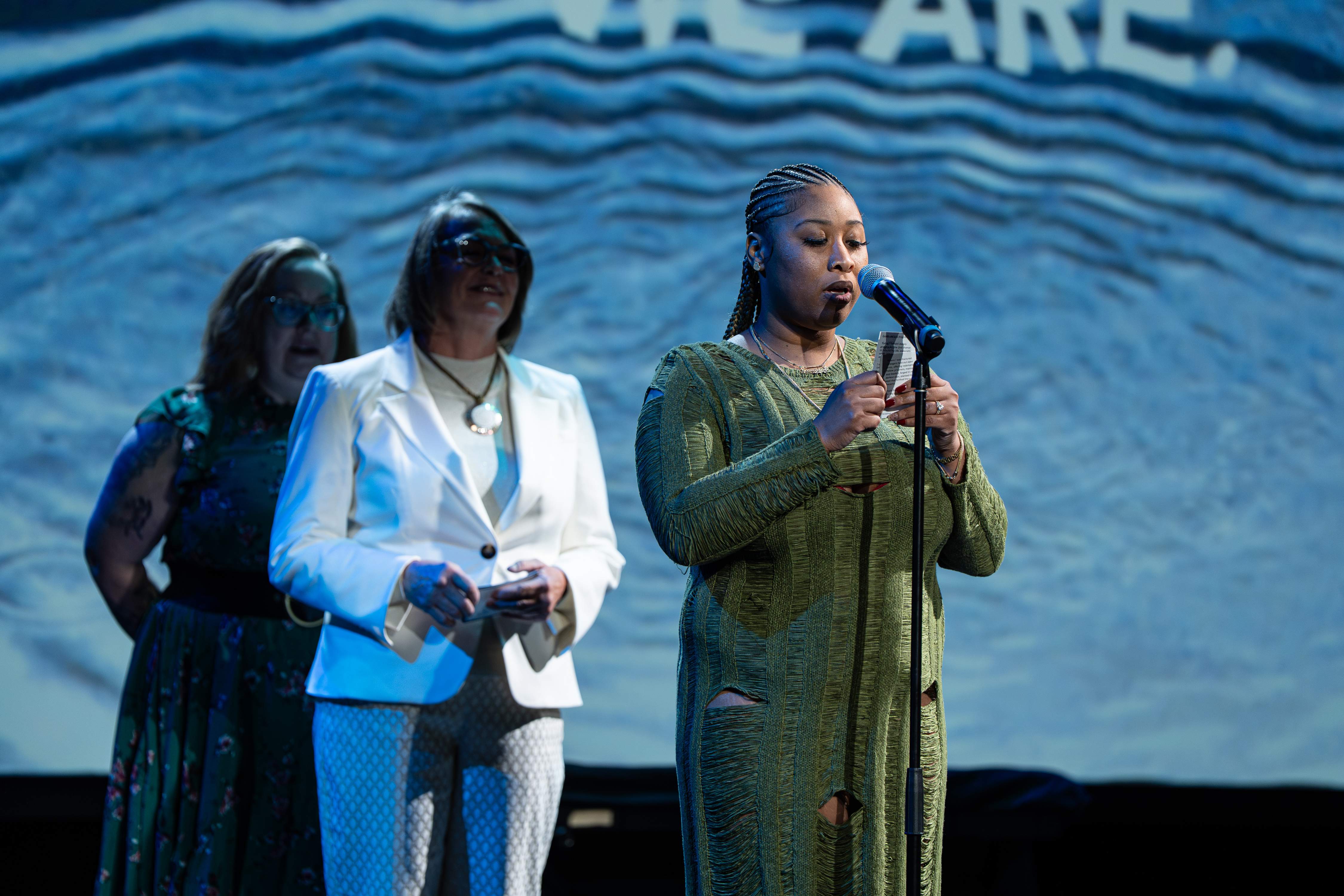 A woman with braided hair, wearing a green textured dress, speaks into a microphone while holding a card, with two women standing behind her.